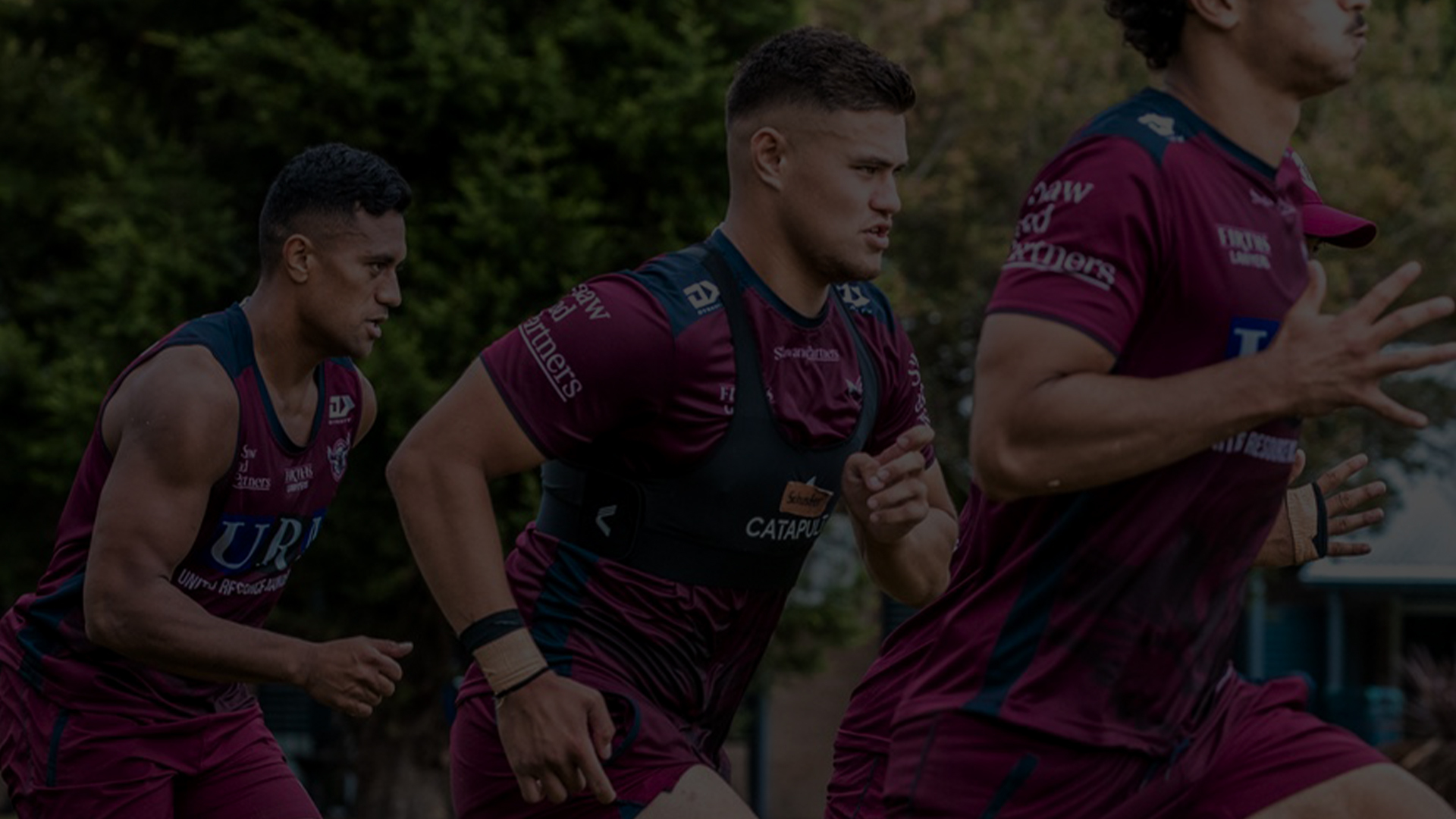 Um grupo de jogadores da NRL é capturado no meio de um intenso exercício de treinamento ao ar livre. Eles estão vestidos com vários tons de camisetas roxas e shorts azuis profundos, enfatizando o trabalho em equipe e o jogo estratégico.