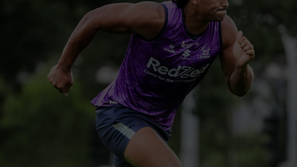 Um jogador da National Rugby League correndo com determinação durante uma sessão de treinamento. Ele está adornado com uma camisa roxa vibrante com logotipos de patrocinadores, combinada com shorts azul marinho. Seu foco e o fundo desfocado transmitem uma sensação de velocidade e agilidade.
