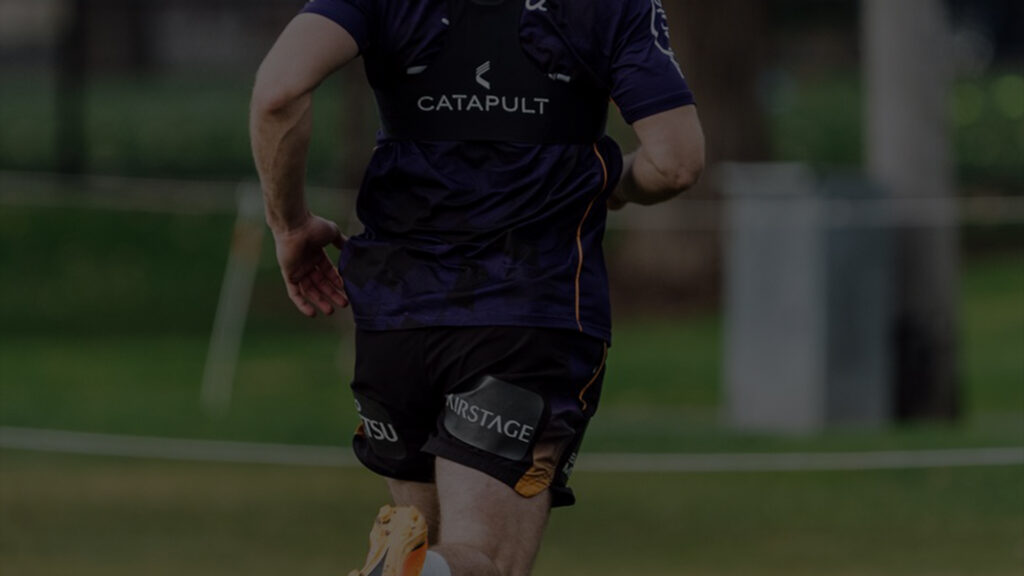 The back view of an NRL player mid-stride on the training field, showcasing the Catapult technology device on his purple and black jersey. The setting is outdoors with a focus on the player's athletic form and the high-tech training equipment.