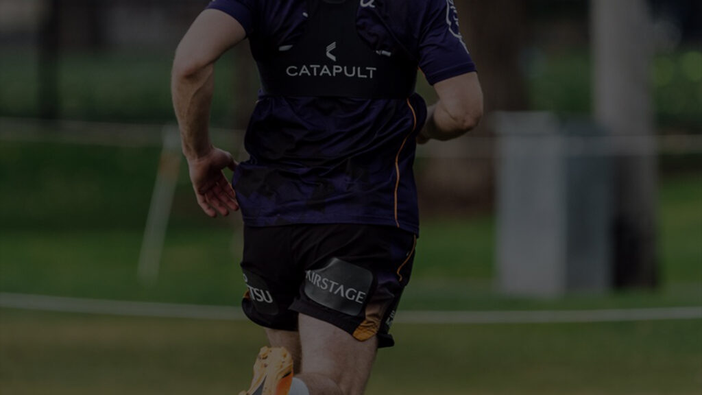 A visão traseira de um jogador da NRL no meio do campo de treinamento, exibindo o dispositivo de tecnologia Catapult em sua camisa roxa e preta. O cenário é ao ar livre com foco na forma atlética do jogador e nos equipamentos de treinamento de alta tecnologia.