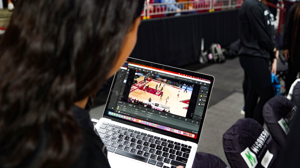 波士顿学院篮球运动员和教练之间正在进行集中互动，笔记本电脑屏幕上可以看到专业视频焦点分析。这凸显了实时视频分析在训练中的整合，这对于提高表现和游戏战术至关重要。