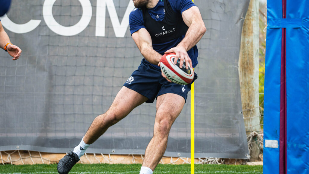 Un jugador de rugby con un chaleco azul oscuro 'CATAPULT' practica pasar el balón con equipo de entrenamiento y un poste de portería al fondo