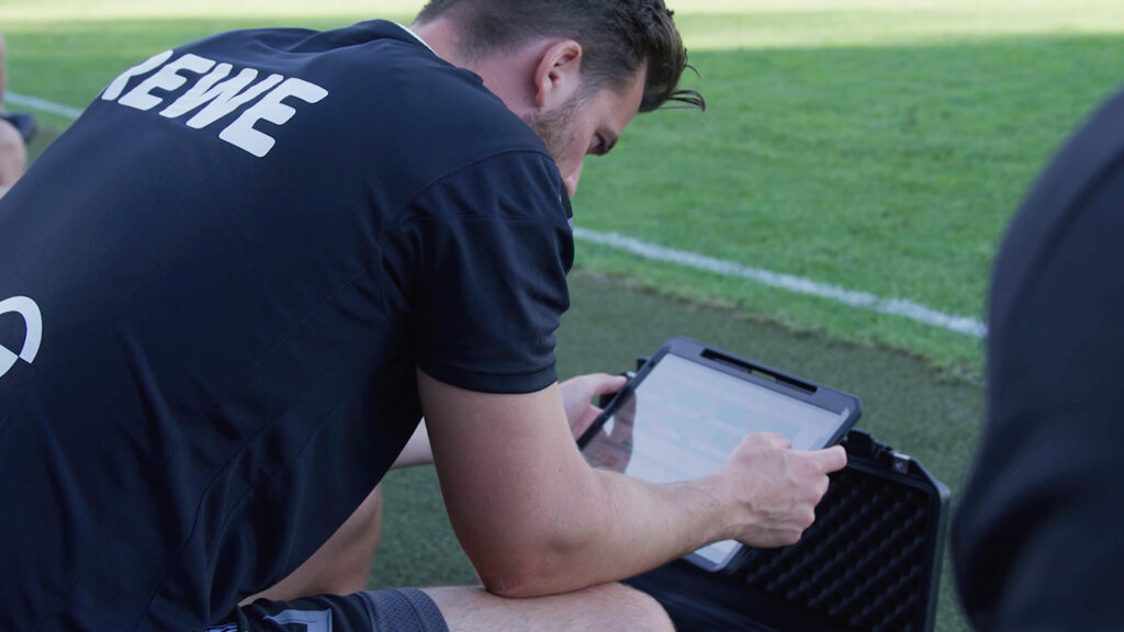 Jogador de futebol de preto analisa dados táticos em um tablet com um treinador durante uma sessão de treinamento, com um campo como pano de fundo