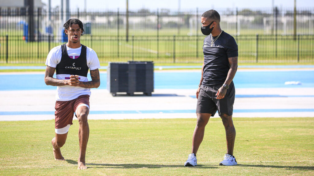 身穿白色和紫色背心、印有“CATAPULT”品牌的男运动员在阳光明媚的田野上训练，黑色教练在一旁观看