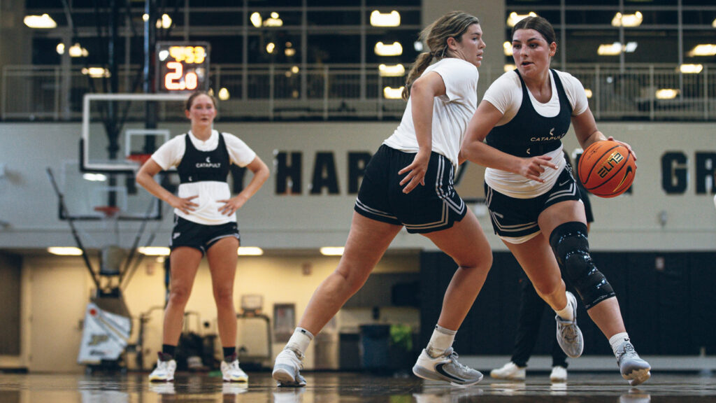 Jogadoras de basquete vestindo coletes 'CATAPULT' participam de um jogo de treinamento indoor, uma no ataque e as outras na defesa