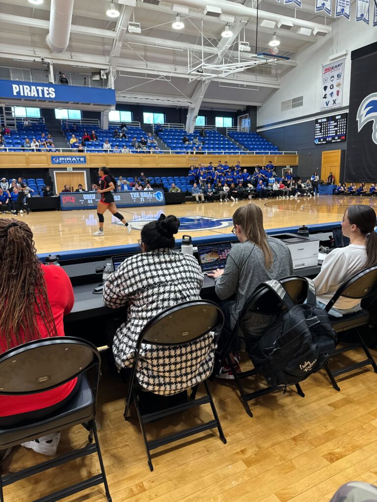 Ein Seitenblick auf einen Basketballplatz, auf dem Analysten und Trainer an einem Tisch sitzen, fleißig an Laptops arbeiten und wahrscheinlich Videoanalysesoftware nutzen, um Strategien zu entwickeln und datengesteuerte Entscheidungen für die Spieler- und Teamentwicklung zu treffen.