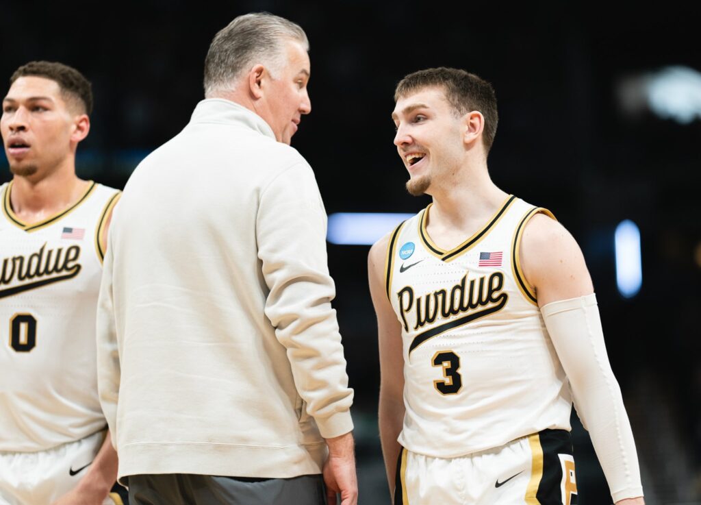 Purdue Boilermakers basketball coach in a beige sweatshirt talking to a smiling player wearing a white and gold uniform with the number 3, on the court side.