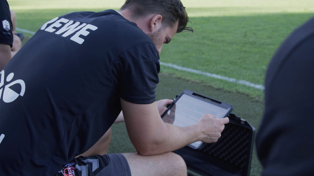 Спортивный аналитик работает на ноутбуке с программным обеспечением для анализа матчей и смотрит на переполненный футбольный стадион из ложи для прессы.