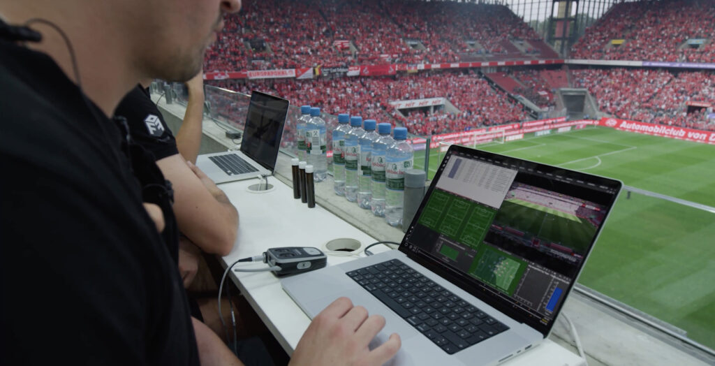 Um analista esportivo trabalhando em um laptop com software de análise de partidas, observando um estádio de futebol lotado da cabine de imprensa.