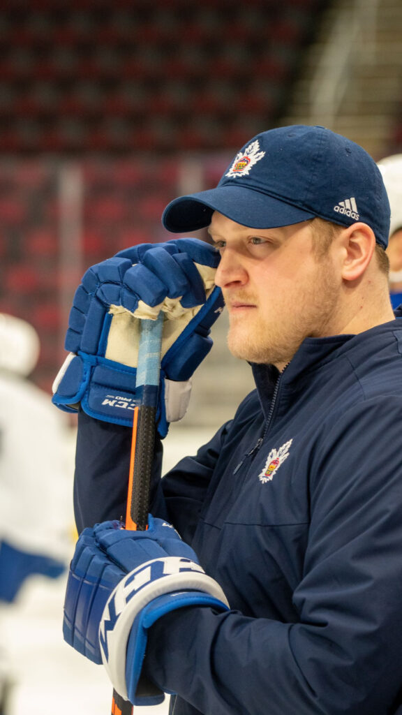 A hockey coach intently watches the ice from the bench during a live game, wearing team apparel.