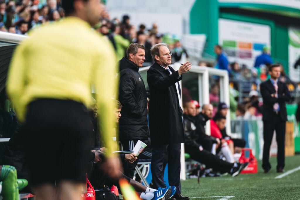 Zwei Trainer gestikulieren und besprechen während eines Fußballspiels an der Seitenlinie die Taktik, im Hintergrund sind Spieler und Schiedsrichter zu sehen.