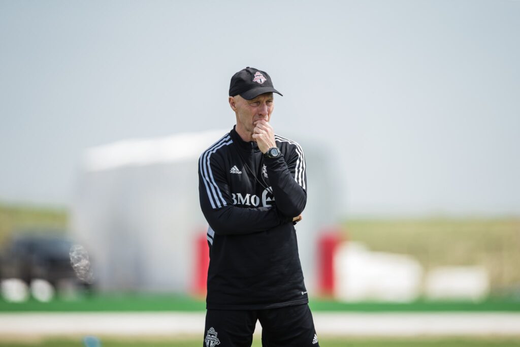 Um treinador, vestido com um agasalho preto do Toronto FC, observa pensativo os jogadores durante um treino.