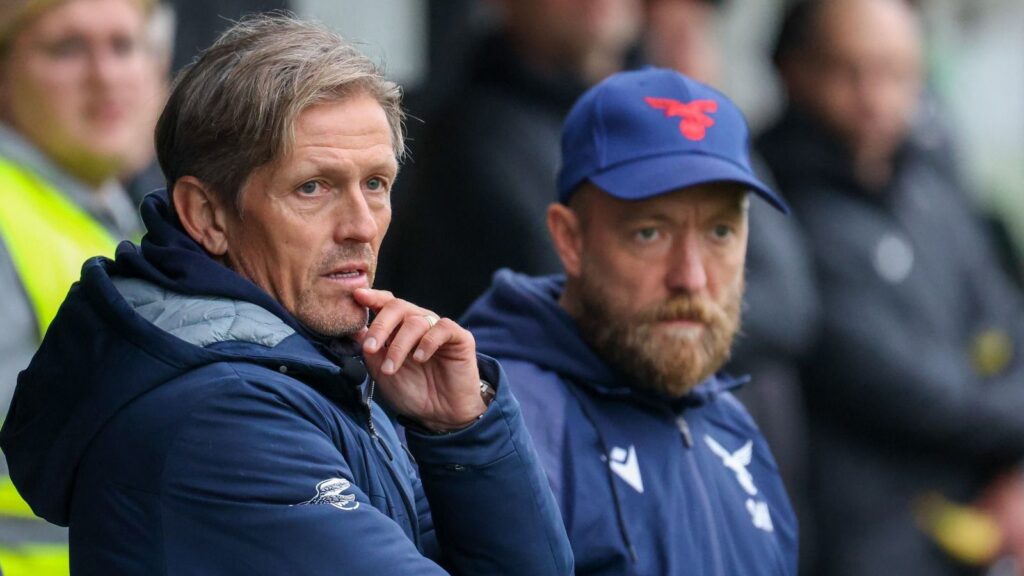 Zwei Fußballtrainer, einer nachdenklich mit dem Finger auf den Lippen, beobachten das Spiel aufmerksam von der Seitenlinie.